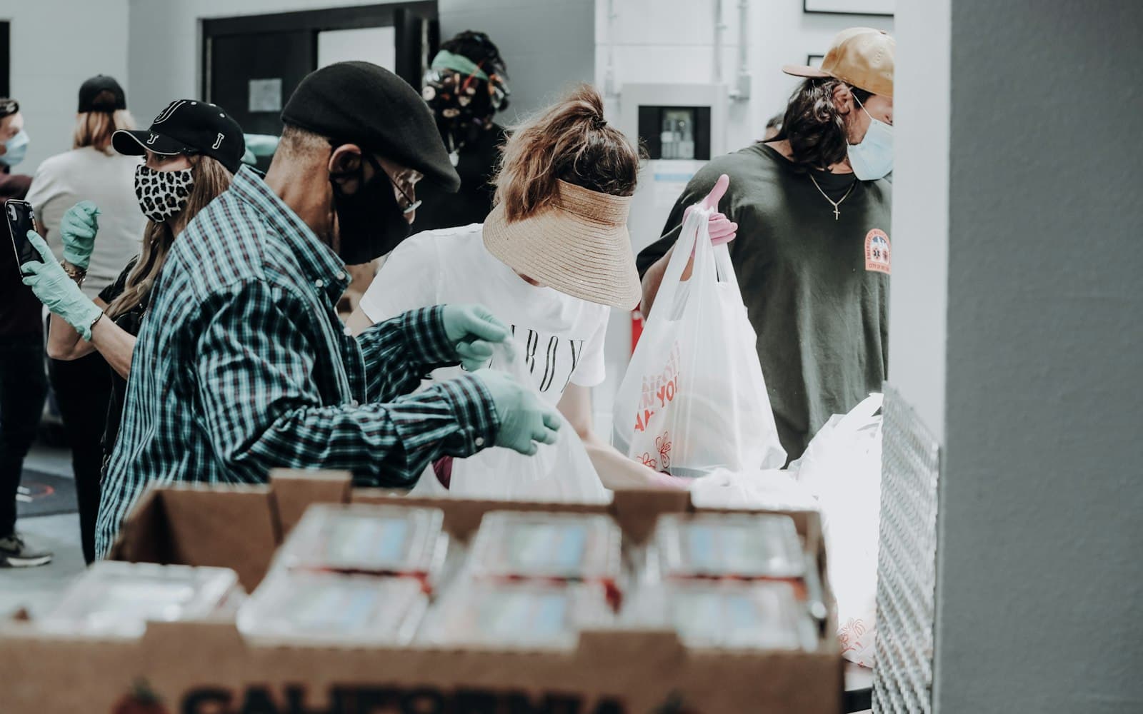 A group of masked volunteers wearing gloves stand around a table sorting bagged groceries, with a box of produce in the foreground.