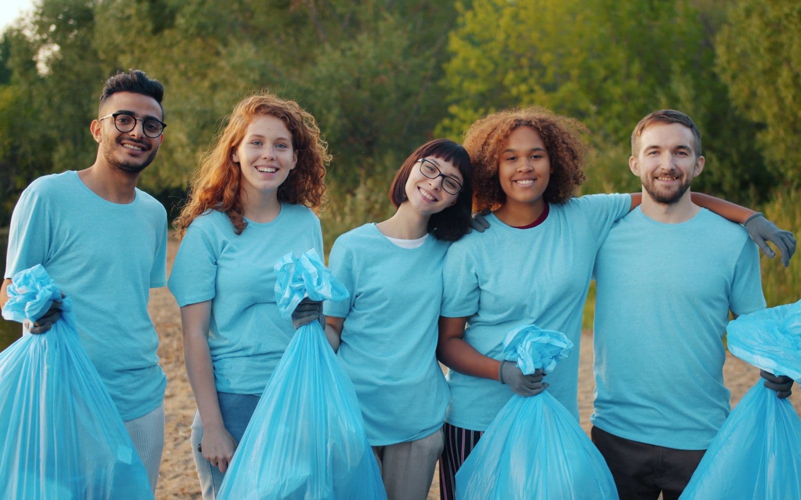 Five smiling volunteers in matching light-blue T-shirts stand arm in arm outdoors near trees, each holding a full trash bag.